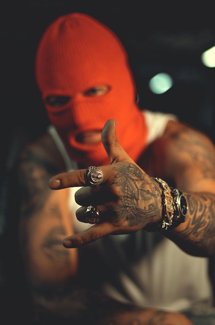 gallery-3 A tattooed man wearing a red ski mask gestures in a dimly lit urban studio.