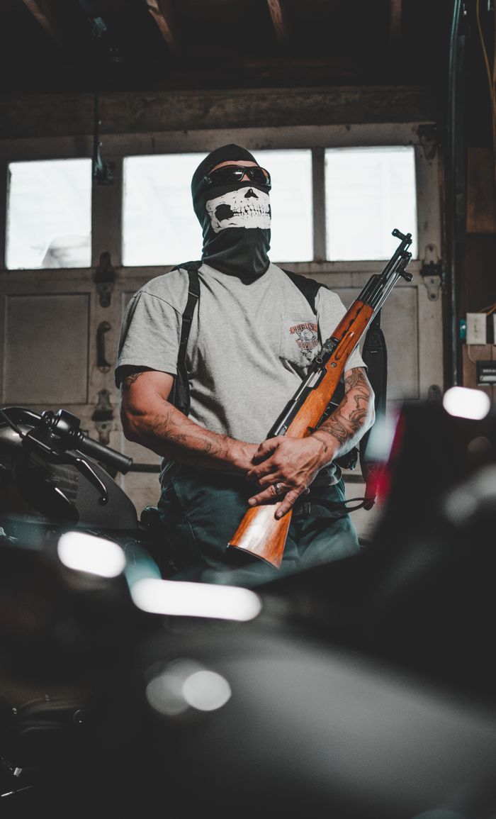 gallery-2 A masked person stands in a garage holding a rifle, surrounded by motorcycles.
